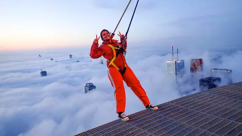 Person in harness leaning backwards over the EdgeWalk. Clouds are below.